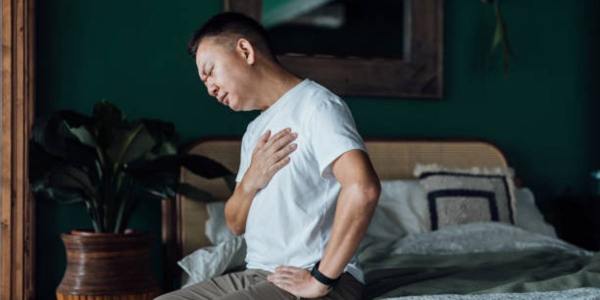 Senior Asian man with eyes closed holding his chest in discomfort, suffering from chest pain while sitting on bed at home. Elderly and health issues concept